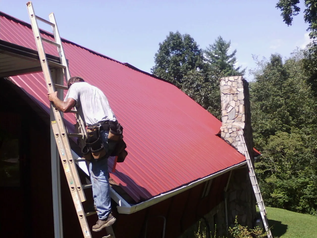 Roofer on a ladder beside a red metal roof and stone chimney — Soffit & Fascia Repair by Roofing Brothers in Halsey